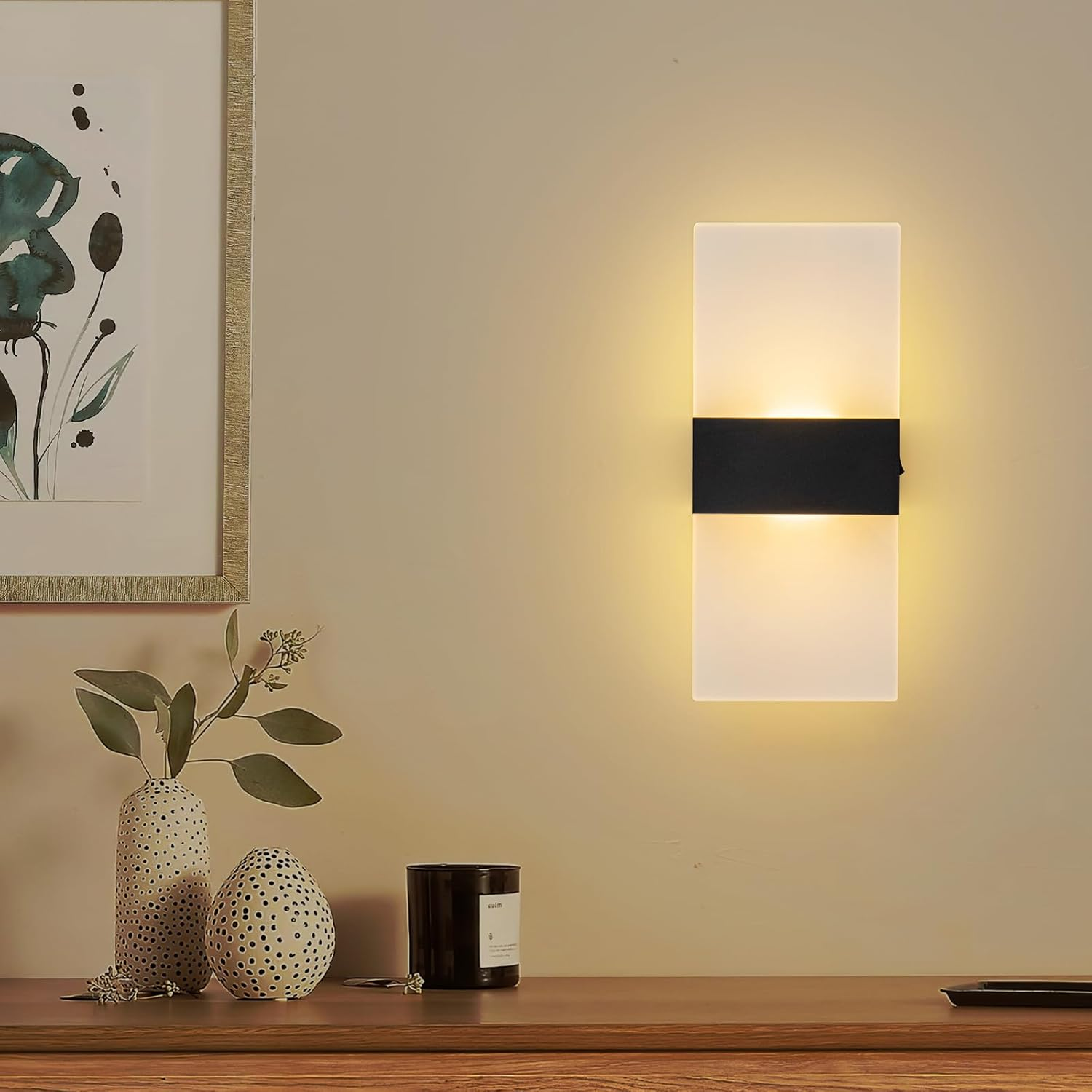 Eine rechteckige Wandleuchte mit schwarzer Mitte leuchtet an einer Wand über Vasen und einer Kerze auf einem Holzregal.