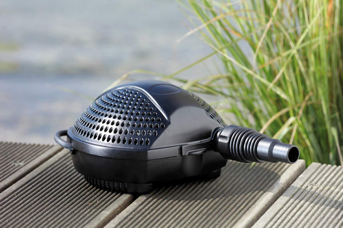 Eine schwarze Teichpumpe mit perforierter Abdeckung und Schlauchanschluss liegt auf Holzplanken vor grünem Gras und Wasser.