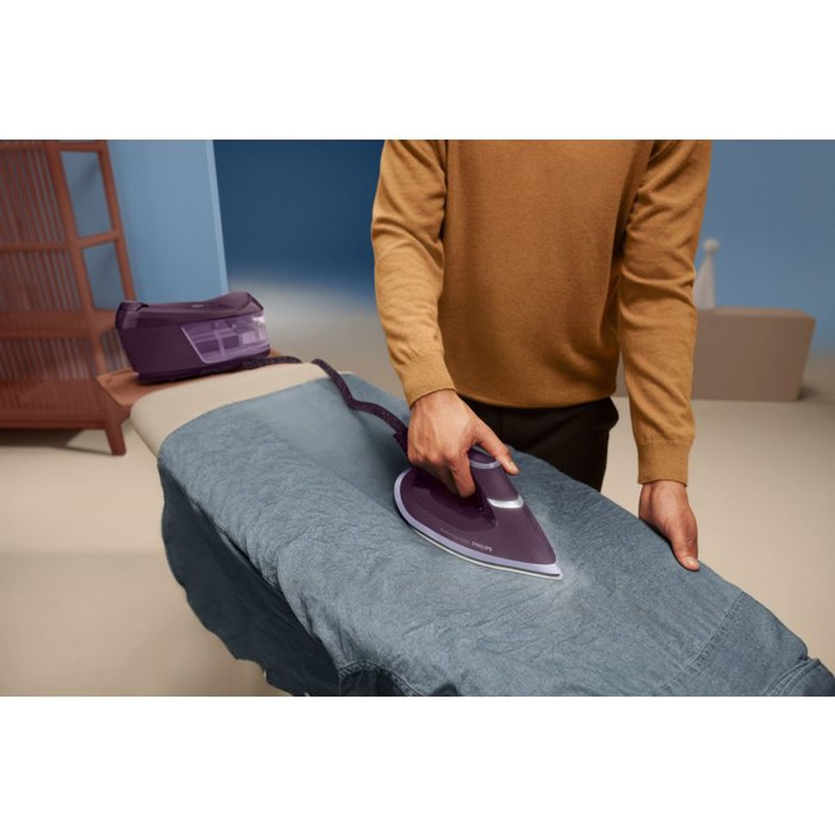 Eine Person bügelt ein graues Hemd mit einem violetten Dampfbügeleisen auf einem Brett vor blauem Hintergrund.