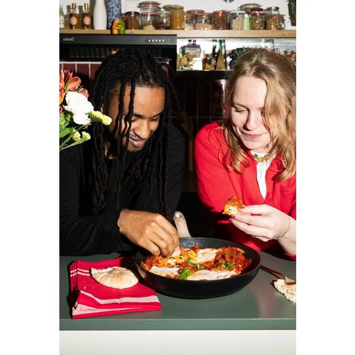 Ein Mann und eine Frau in einer Küche essen Shakshuka aus einer Pfanne mit Fladenbrot.