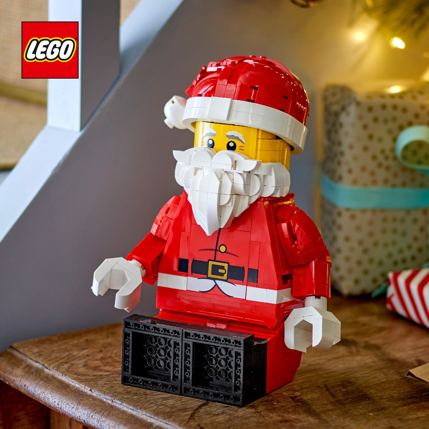 Figurka Świętego Mikołaja z Lego siedzi na drewnianym stole, a w tle widać prezenty i logo Lego.
