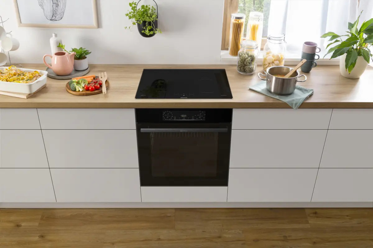 Moderne Küche mit schwarzem Induktionskochfeld, schwarzem Backofen in weißem Schrank, Lebensmitteln und Pflanzen.