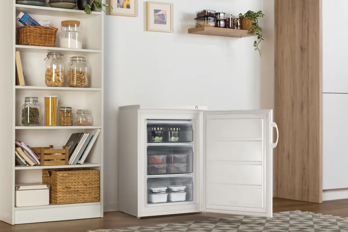 Ein kleiner weißer Gefrierschrank mit offener Tür steht neben einem Regal mit Gläsern und Körben.