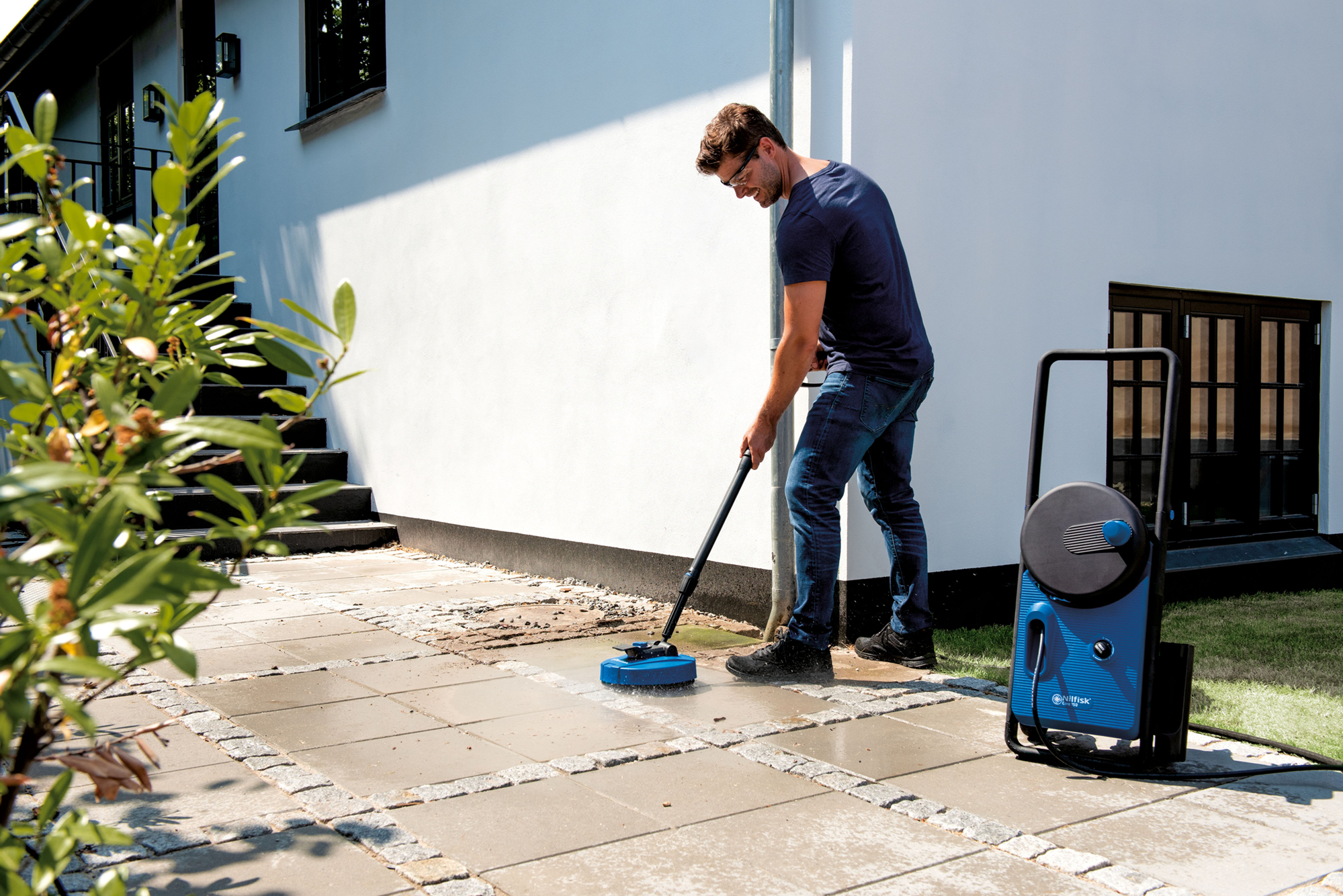 Un uomo pulisce le piastrelle grises del patio in giardino usando un pulisci superfici blu e un'idropulitrice.