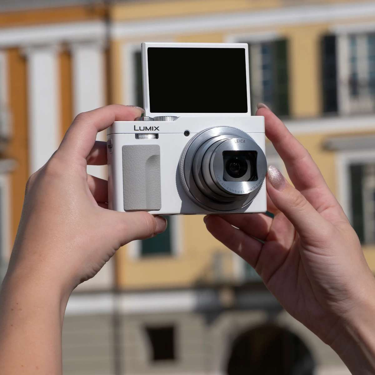 Dwie dłonie trzymają biały aparat Lumix z podniesionym ekranem przed rozmytą fasadą.