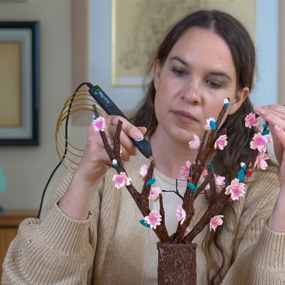 Una mujer usa un bolígrafo 3D para unir flores rosas a ramas en un florero.