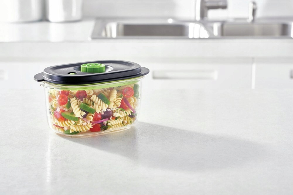 Un recipiente transparente con ensalada de pasta en una encimera de cocina, con fregadero y muebles al fondo.