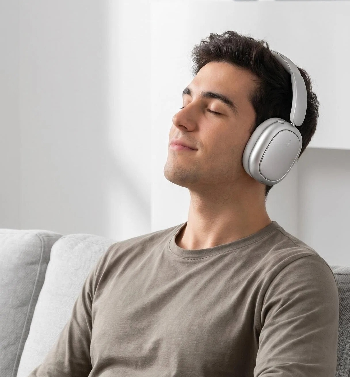 Un hombre con cabello oscuro usa auriculares blancos y está sentado en un sofá gris con los ojos cerrados.