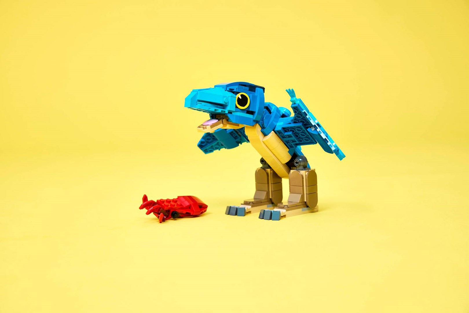 Niebieski dinozaur z klocków Lego stoi przed małym czerwonym robakiem na żółtym tle.