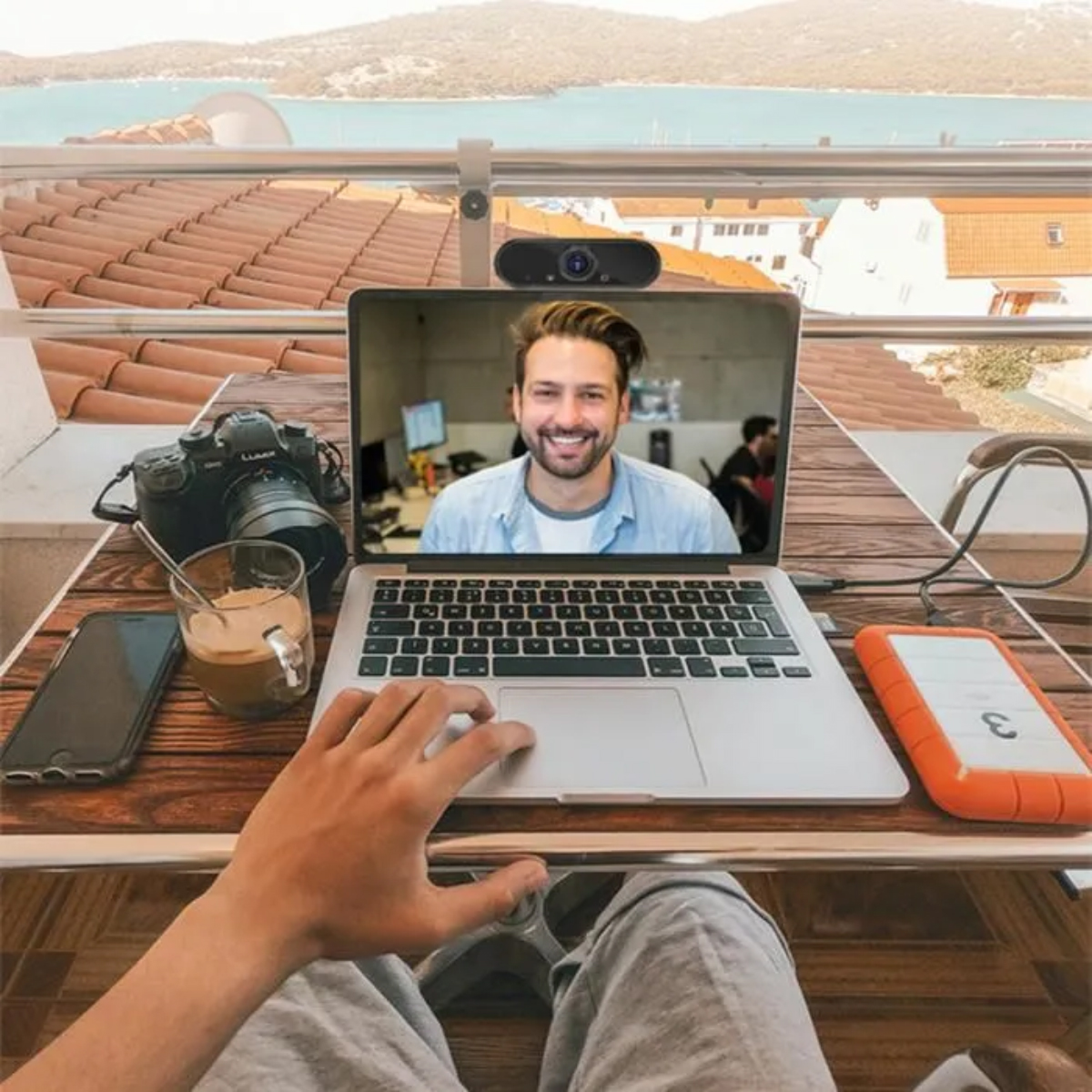 Ein Laptop mit Videoanruf, Kamera, Kaffeetasse und Smartphone auf einem Tisch mit Blick auf das Meer und Dächer.