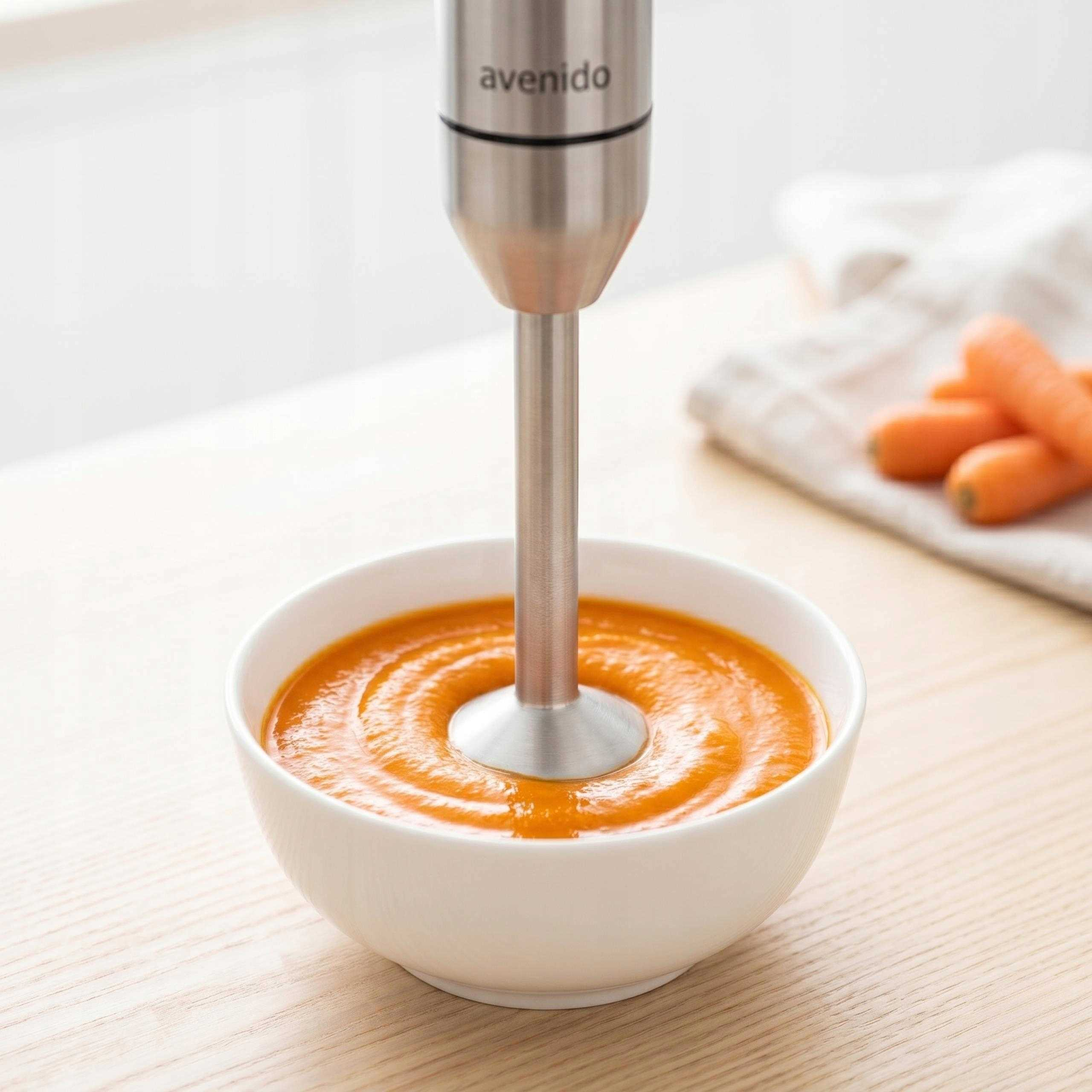 Blender ręczny miksuje pomarańczową zupę w białej misce na drewnianym stole, w tle widoczne marchewki.