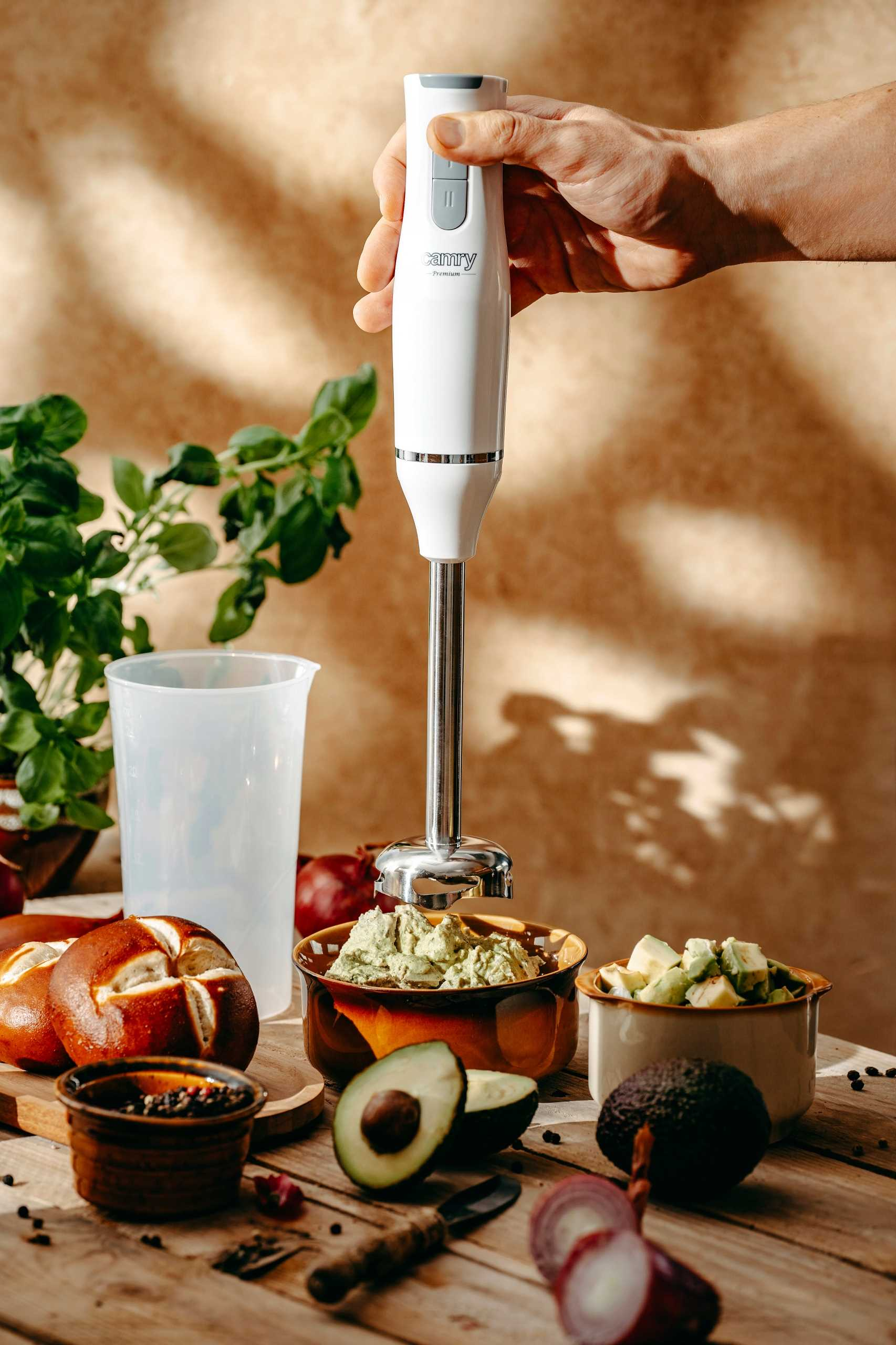 Ręka trzyma biały blender ręczny nad miską guacamole na drewnianym stole z awokado i chlebem.