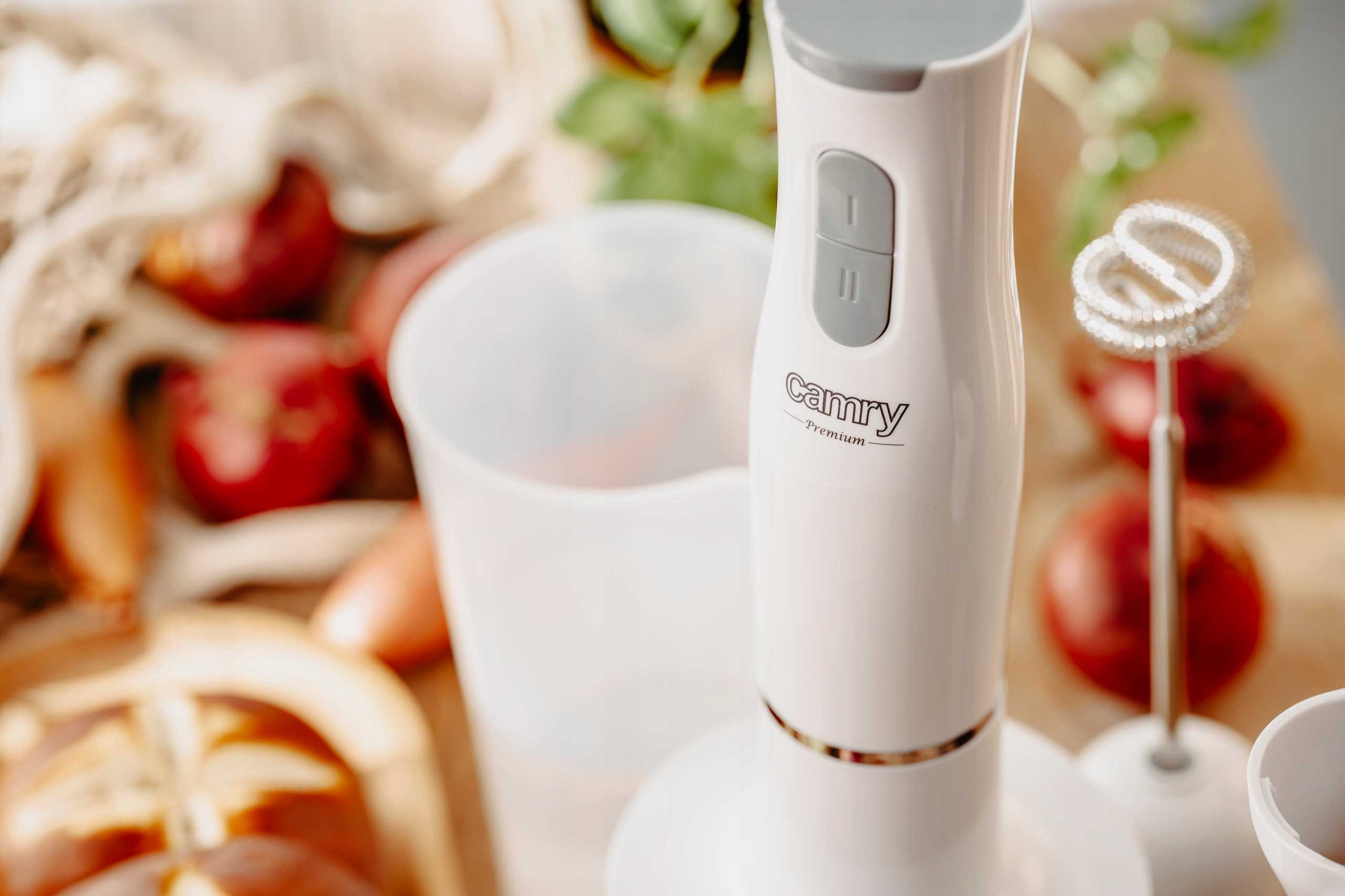 Biały blender ręczny Camry z szarymi przyciskami stoi na drewnianym stole, w otoczeniu jedzenia.