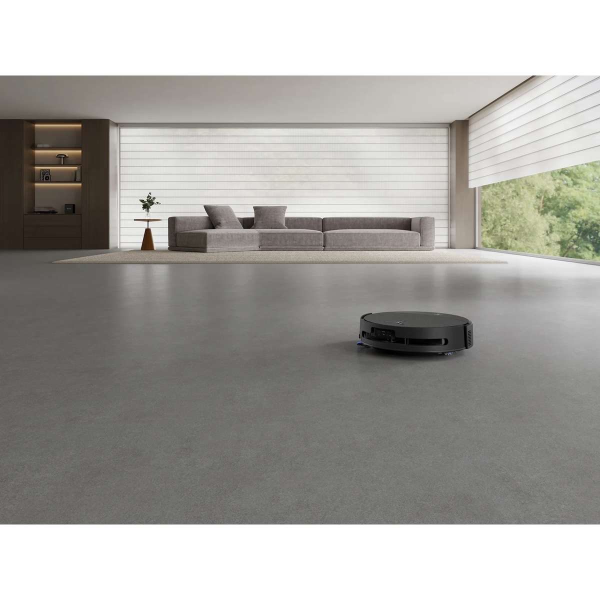 Ein schwarzer Saugroboter steht auf einem grauen Boden in einem minimalistischen Wohnzimmer mit grauem Sofa.
