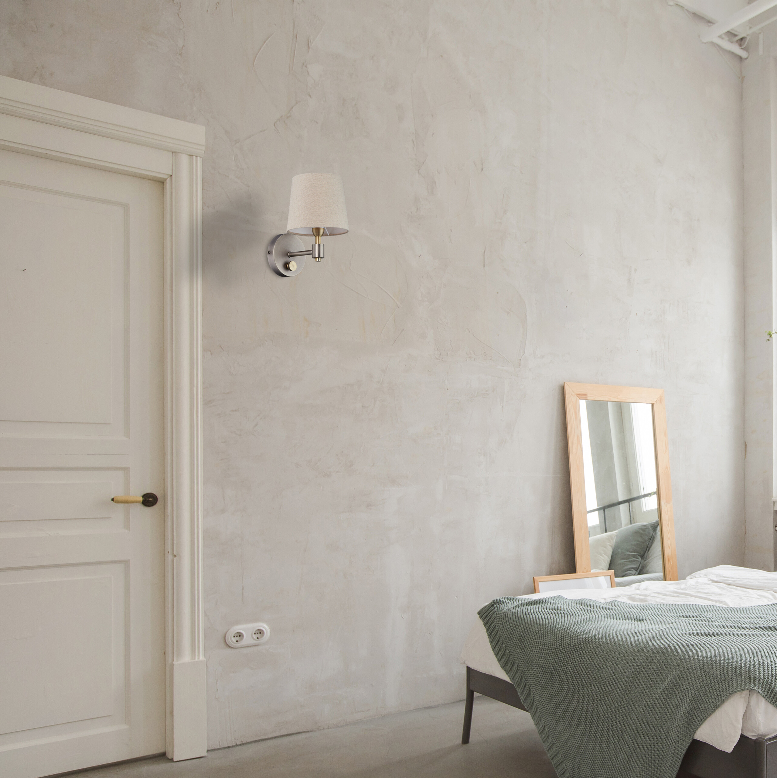 Eine beigefarbene Wandlampe ist an einer strukturierten grauen Wand über einem Bett und einem Spiegel montiert.