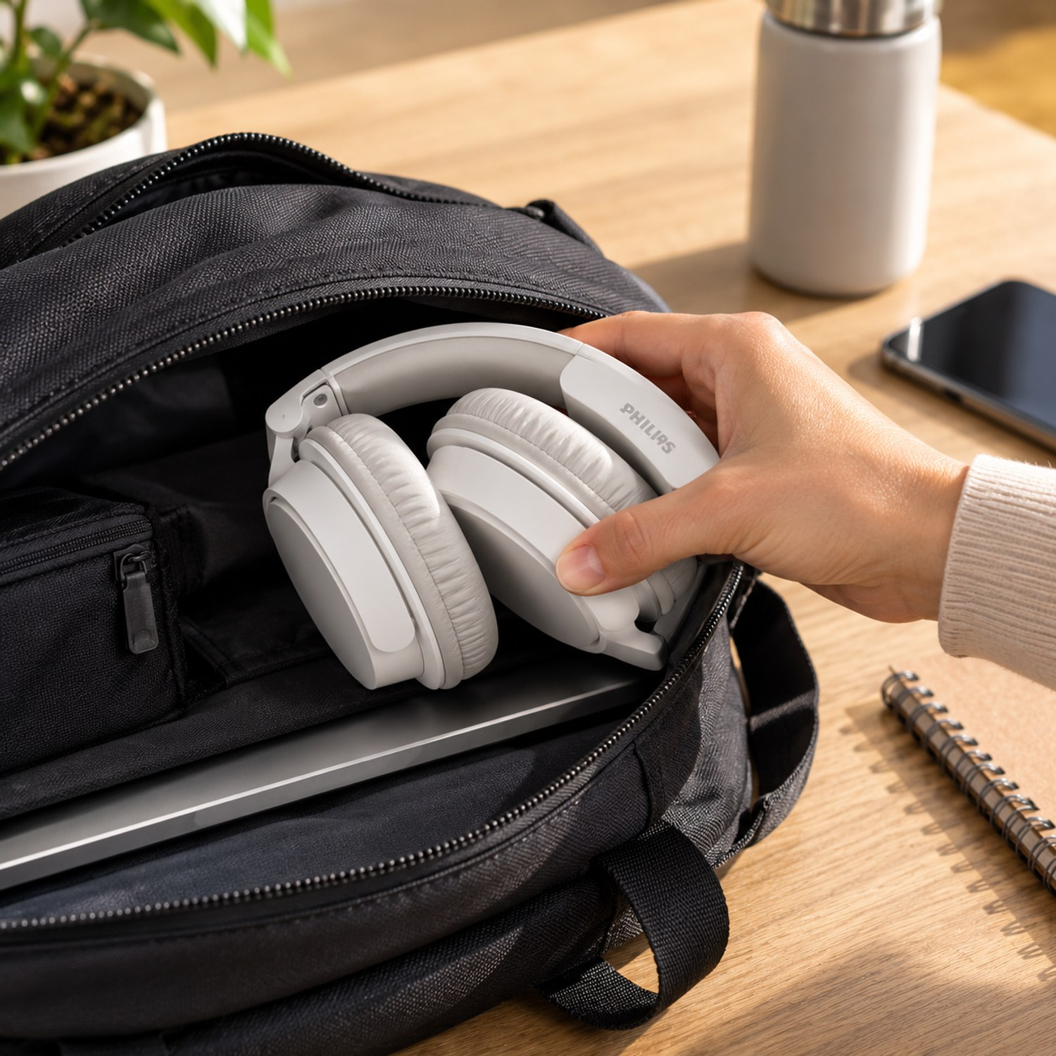 Eine Hand legt weiße Philips Kopfhörer in eine offene schwarze Tasche auf einem Holztisch.