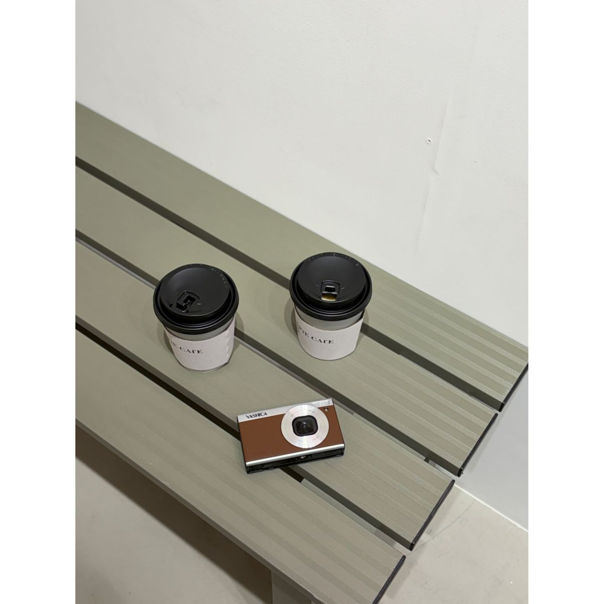 Zwei Pappbecher und eine kleine braun-silberne Kamera liegen auf einer grünen Holzbank vor einer weißen Wand.