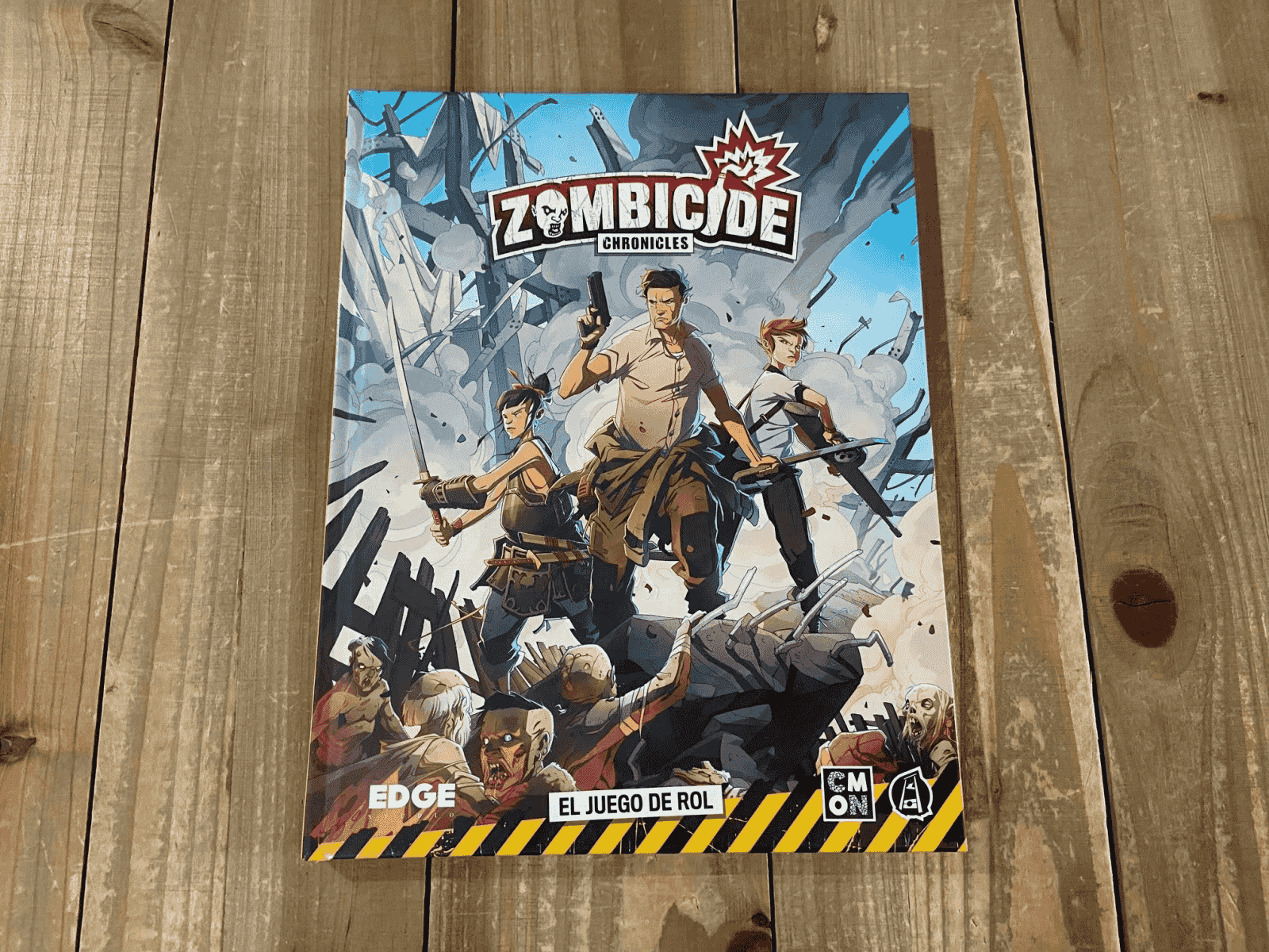 Libro de rol Zombicide Chronicles con tres héroes y zombis de fondo sobre una mesa de madera.