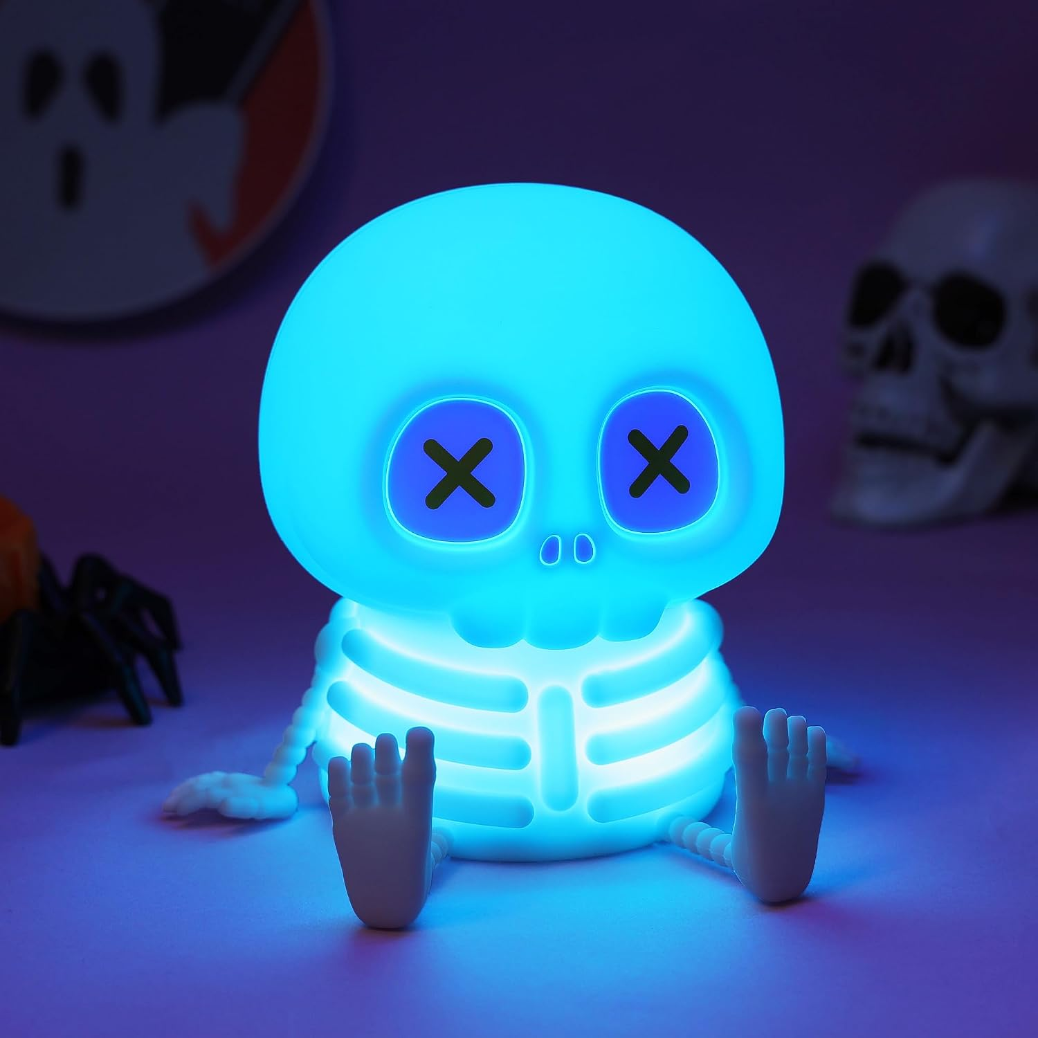 Ein leuchtend blaues Skelett-Nachtlicht mit X-Augen sitzt auf einer violetten Oberfläche mit Halloween-Dekoration.