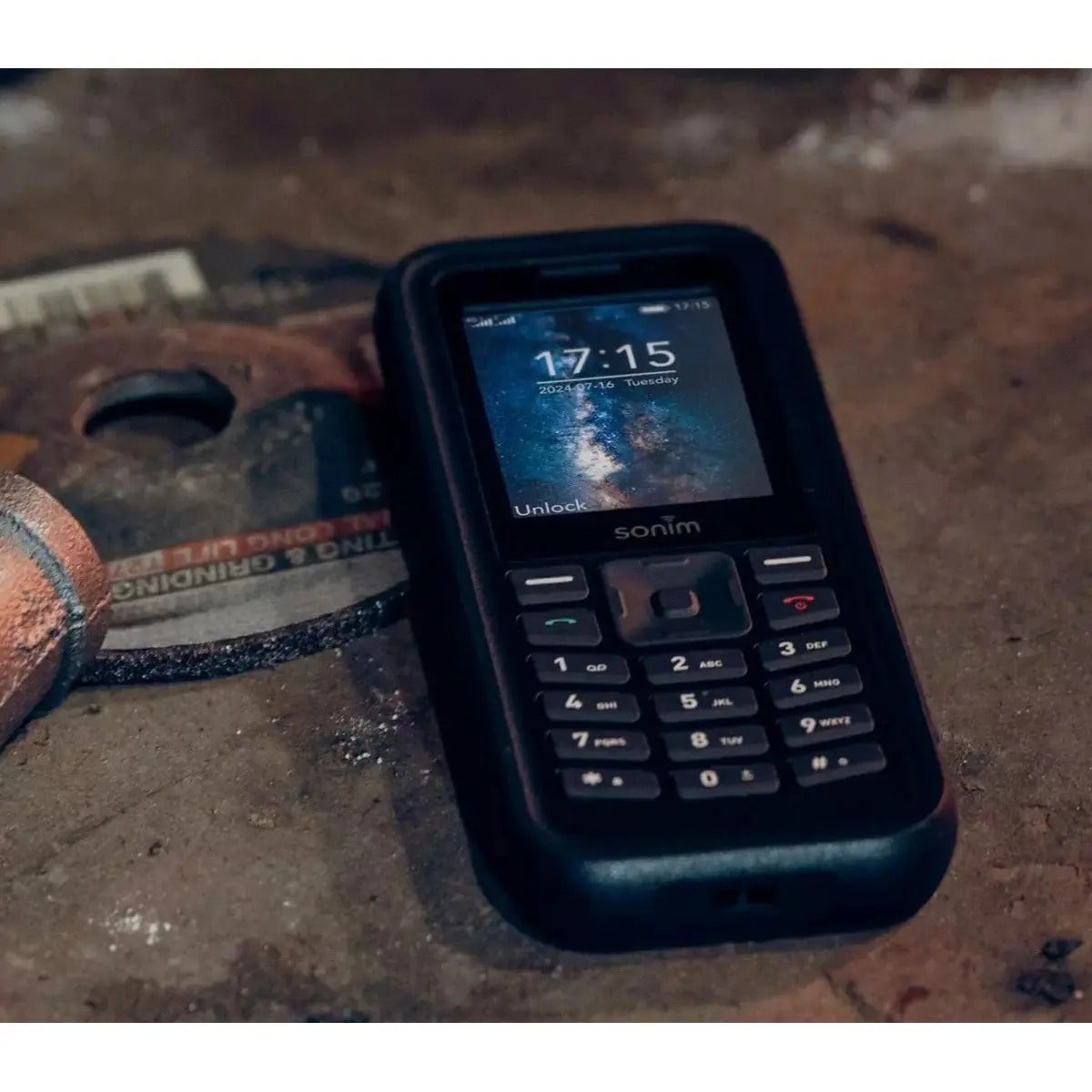 Un teléfono resistente Sonim descansa sobre una superficie metálica rugosa junto a piezas de herramientas.