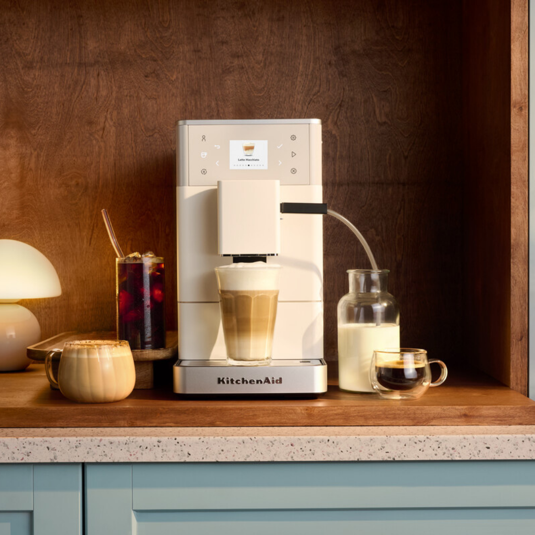 Ahşap tezgah üzerinde latte, buzlu kahve, süt ve fincanların bulunduğu bej renkli KitchenAid kahve makinesi.