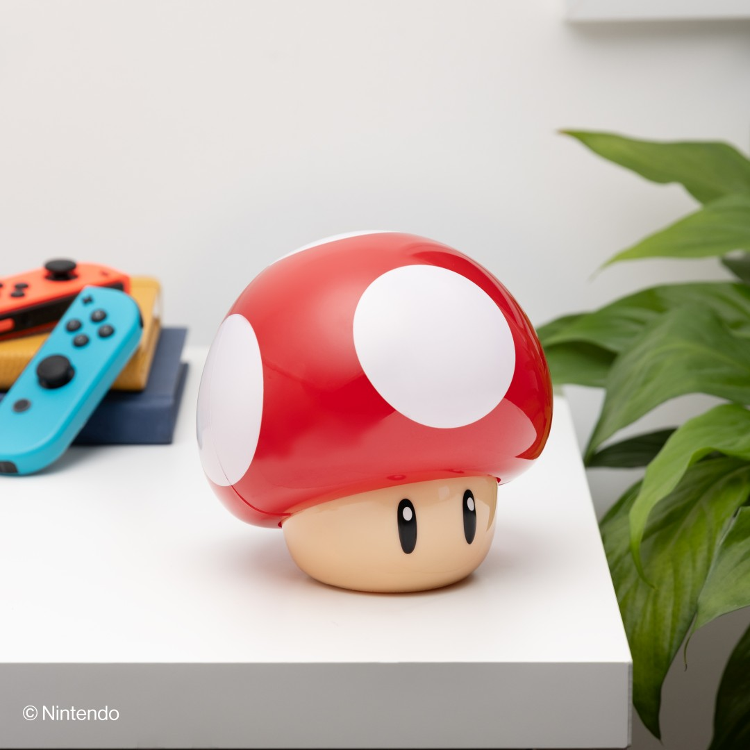 Eine rote Super Mario Pilzlampe steht auf einem weißen Schreibtisch neben Büchern, Controllern und einer Pflanze.