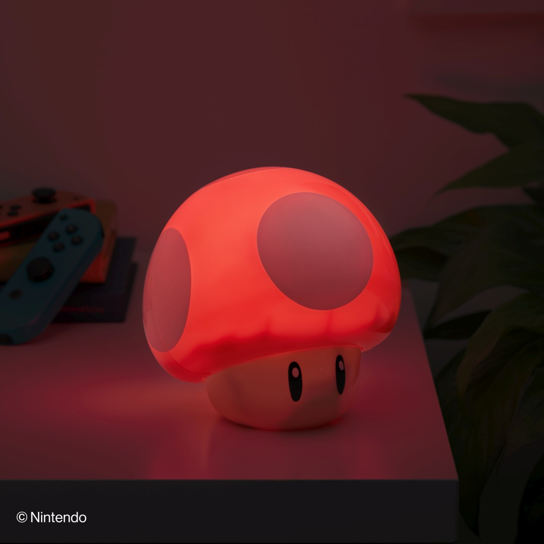 Eine leuchtende rote Super Mario Pilz-Lampe steht auf einem Tisch neben Videospielreglern und Pflanzen.