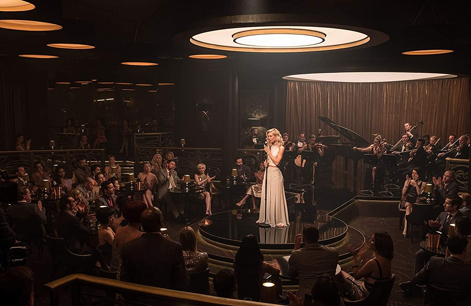 Una cantante rubia actúa en un escenario redondo en un club elegante y oscuro, rodeada de público y músicos.