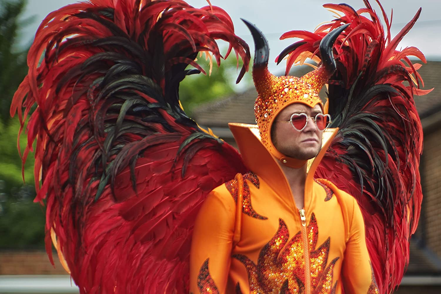 Un hombre disfrazado de diablo con plumas rojas y negras, grandes cuernos y gafas de sol en forma de corazón.