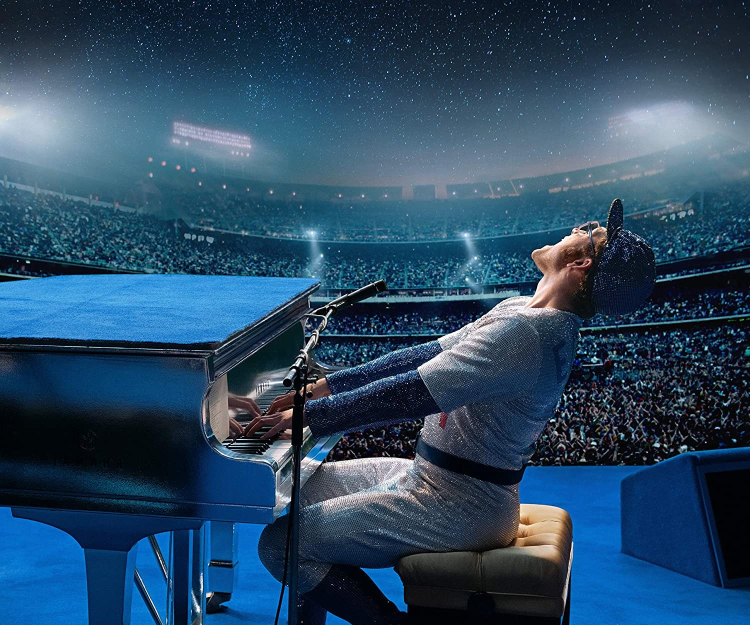 Un músico con ropa brillante toca el piano en un estadio lleno bajo un cielo estrellado.