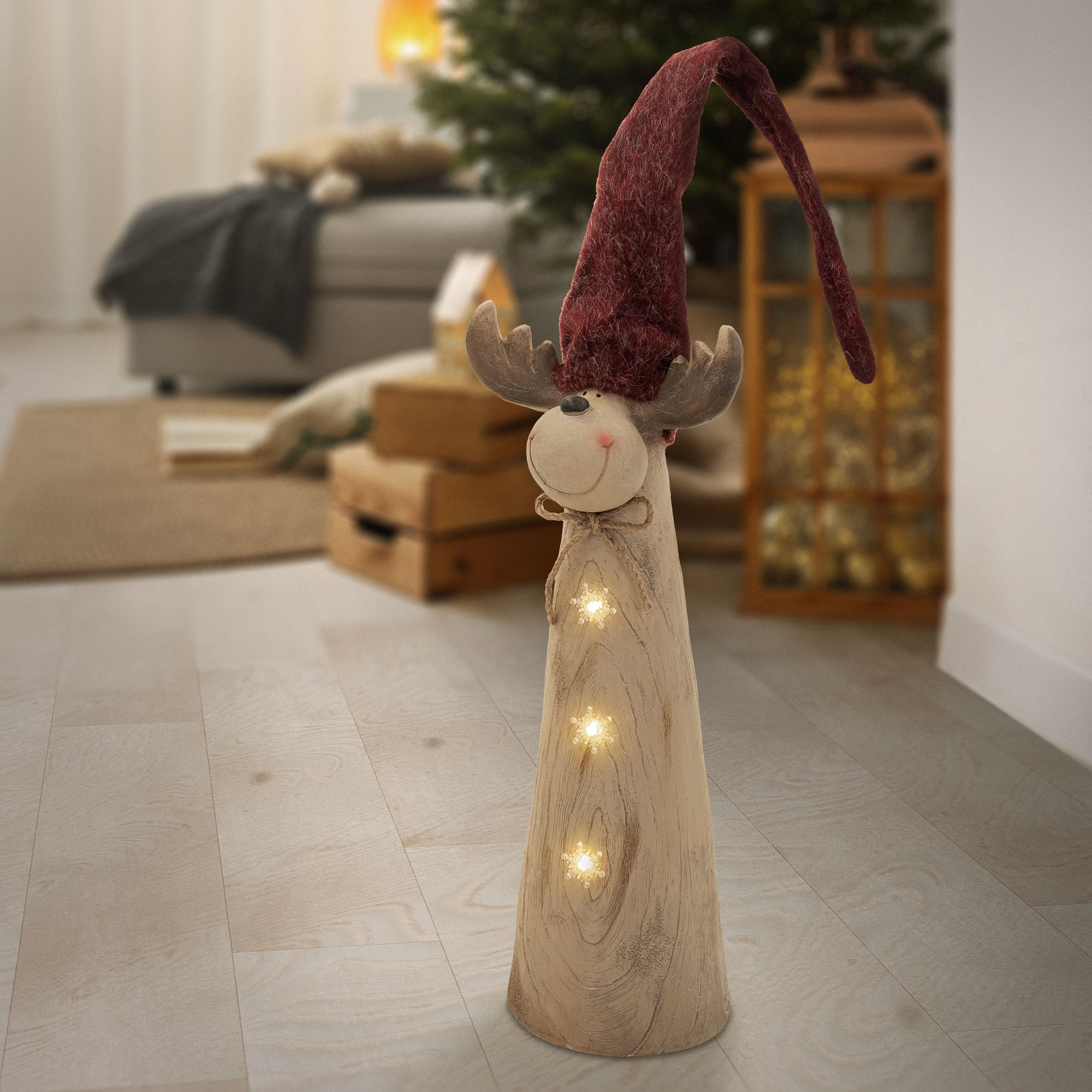 Eine hölzerne Rentierfigur mit roter Mütze und Lichtern steht auf einem Boden, im Hintergrund ist ein Wohnzimmer.