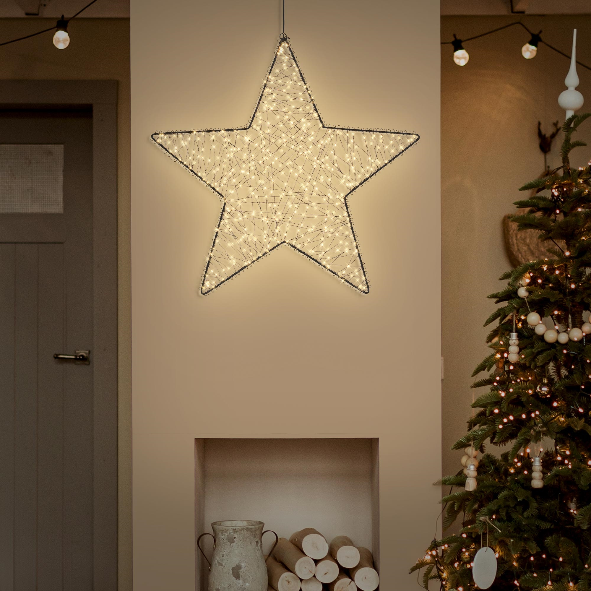 Eine beleuchtete Drahtstern-Dekoration an der Wand über einem Kamin, neben einem geschmückten Weihnachtsbaum.
