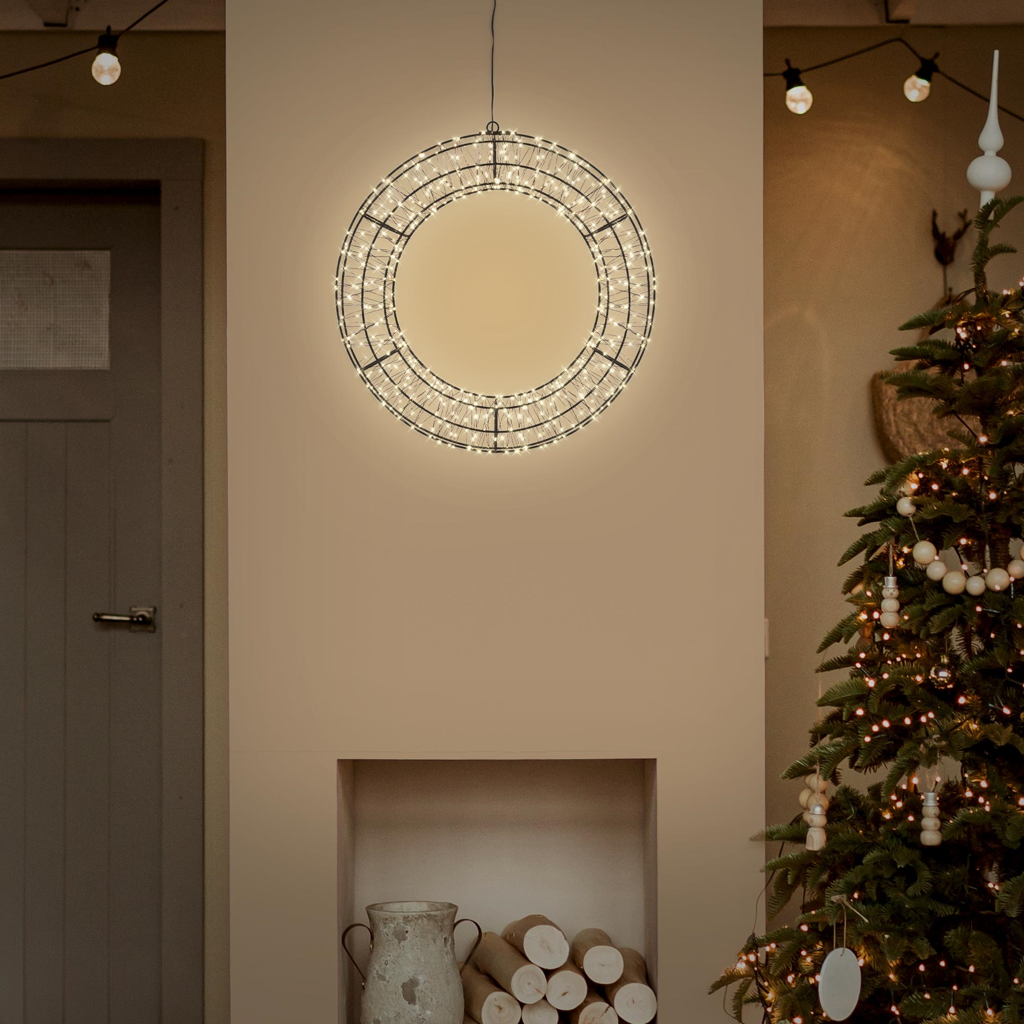 Beleuchteter Metallring als Kranz hängt an der Wand, daneben ein geschmückter Weihnachtsbaum.