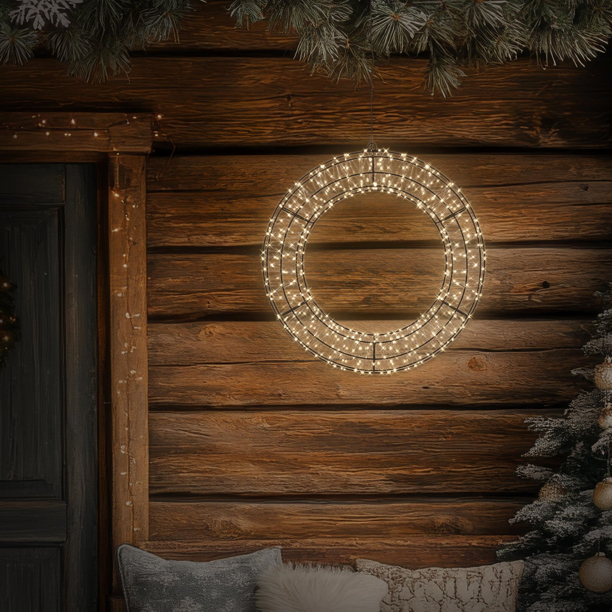 Beleuchteter Lichterkranz hängt vor Holzwand einer Hütte mit Tür und Tannenbaum.