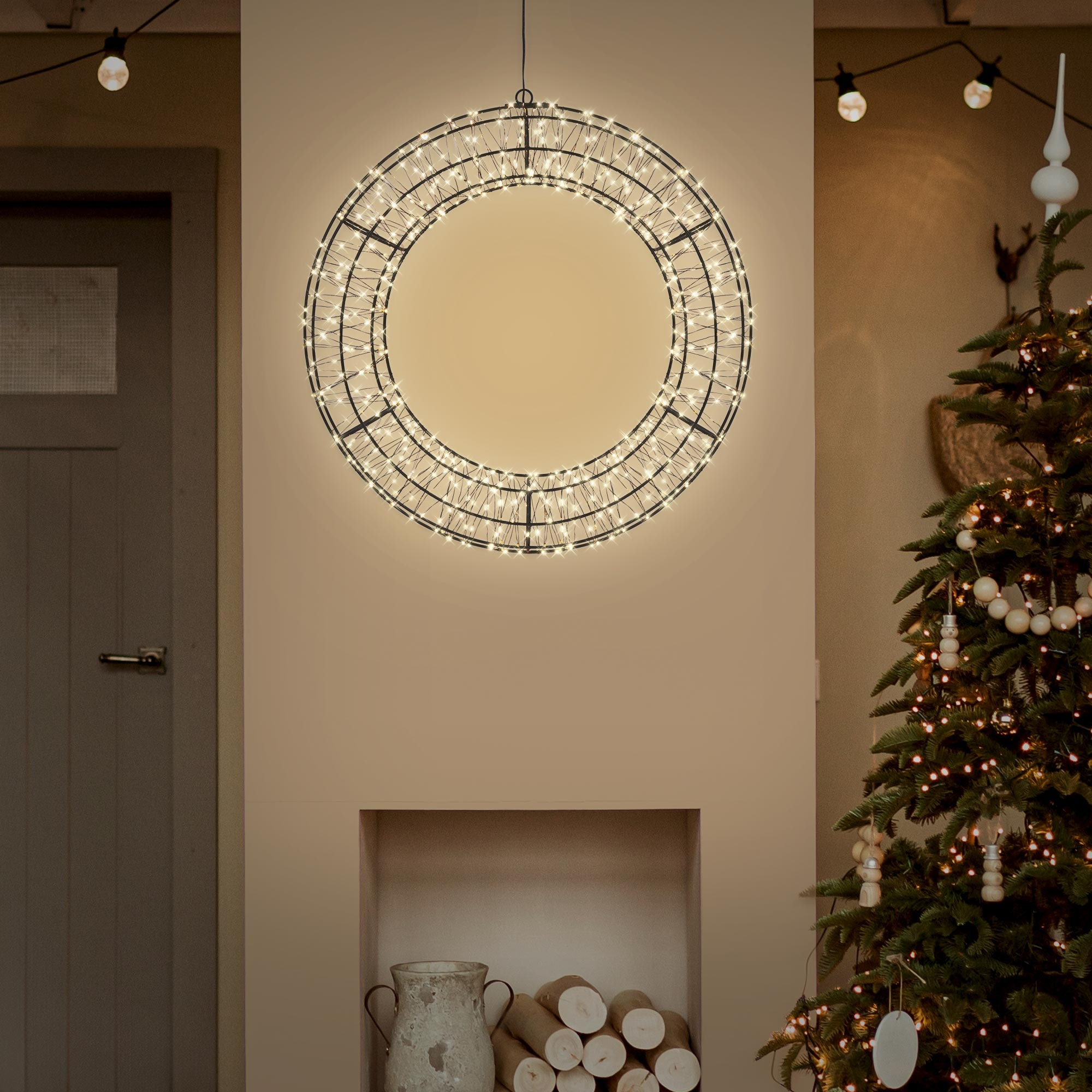 Ein großer, runder, beleuchteter Weihnachtskranz hängt vor einem neutralen Wandhintergrund neben einem Weihnachtsbaum.