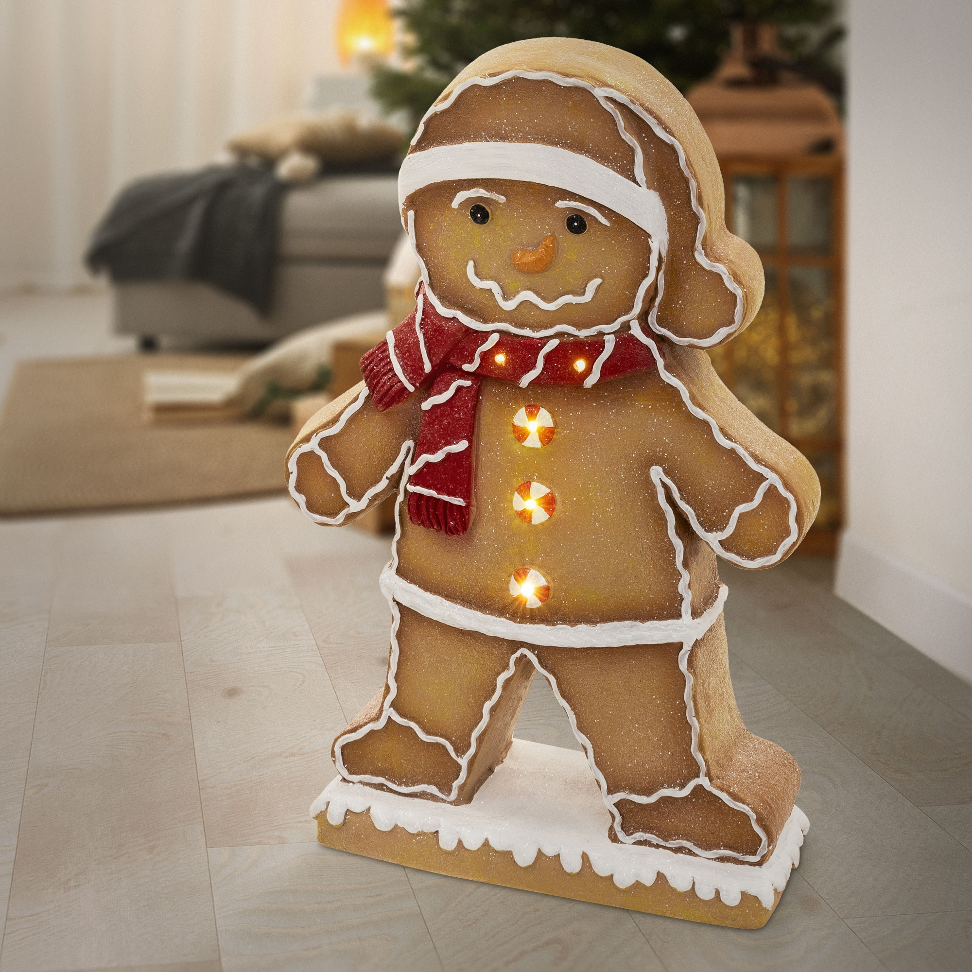 Eine leuchtende Lebkuchenmann-Figur mit rotem Schal und Mütze steht auf einem Holzboden in einem Wohnzimmer.