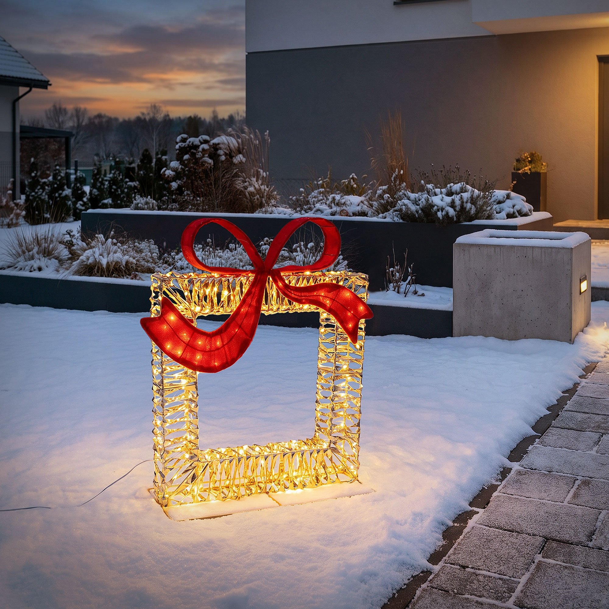 Beleuchtete Geschenkskulptur mit roter Schleife im Schnee vor einem modernen Haus.
