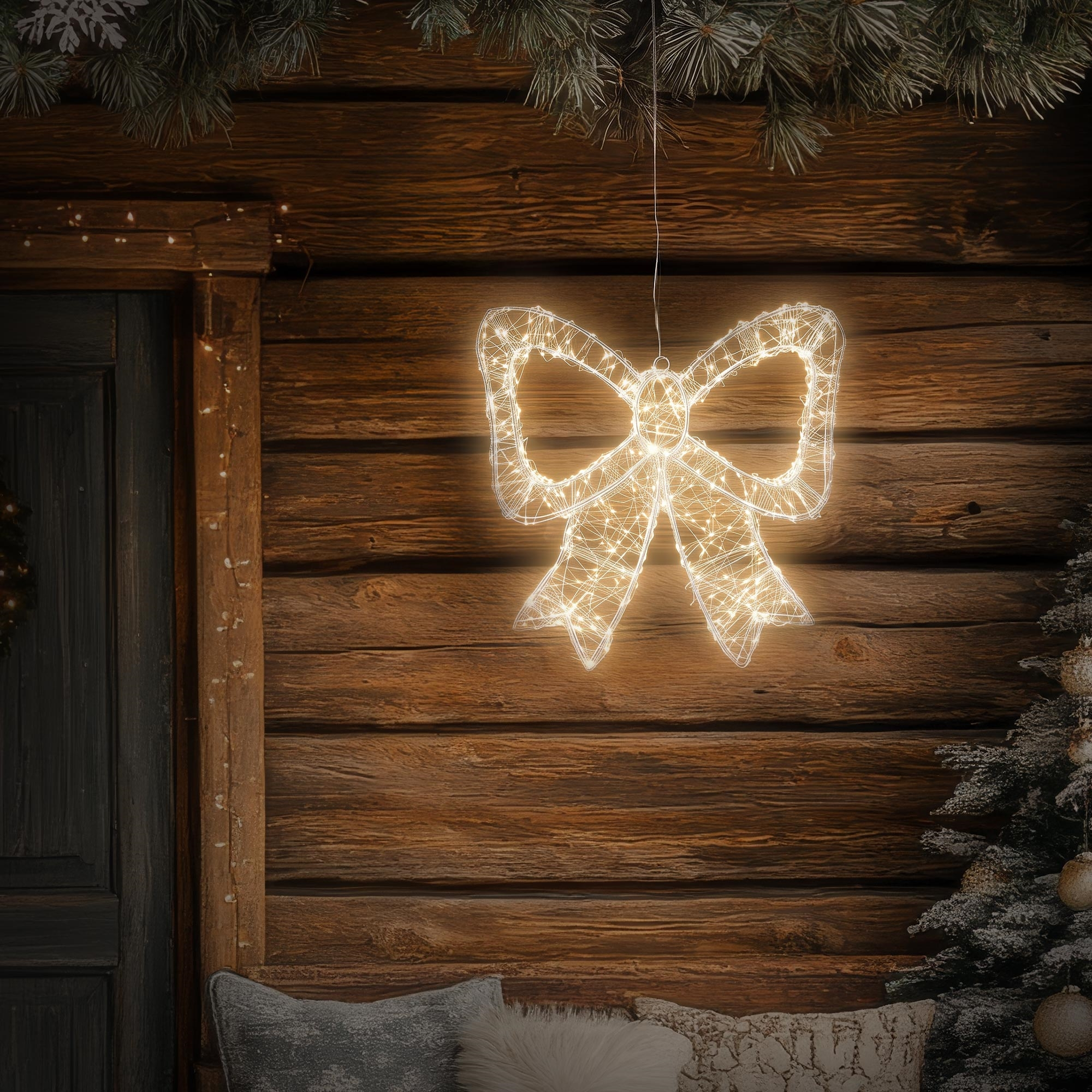 Eine leuchtende Weihnachtsmasche hängt vor einer Holzwand mit Tannengrün und Kissen.