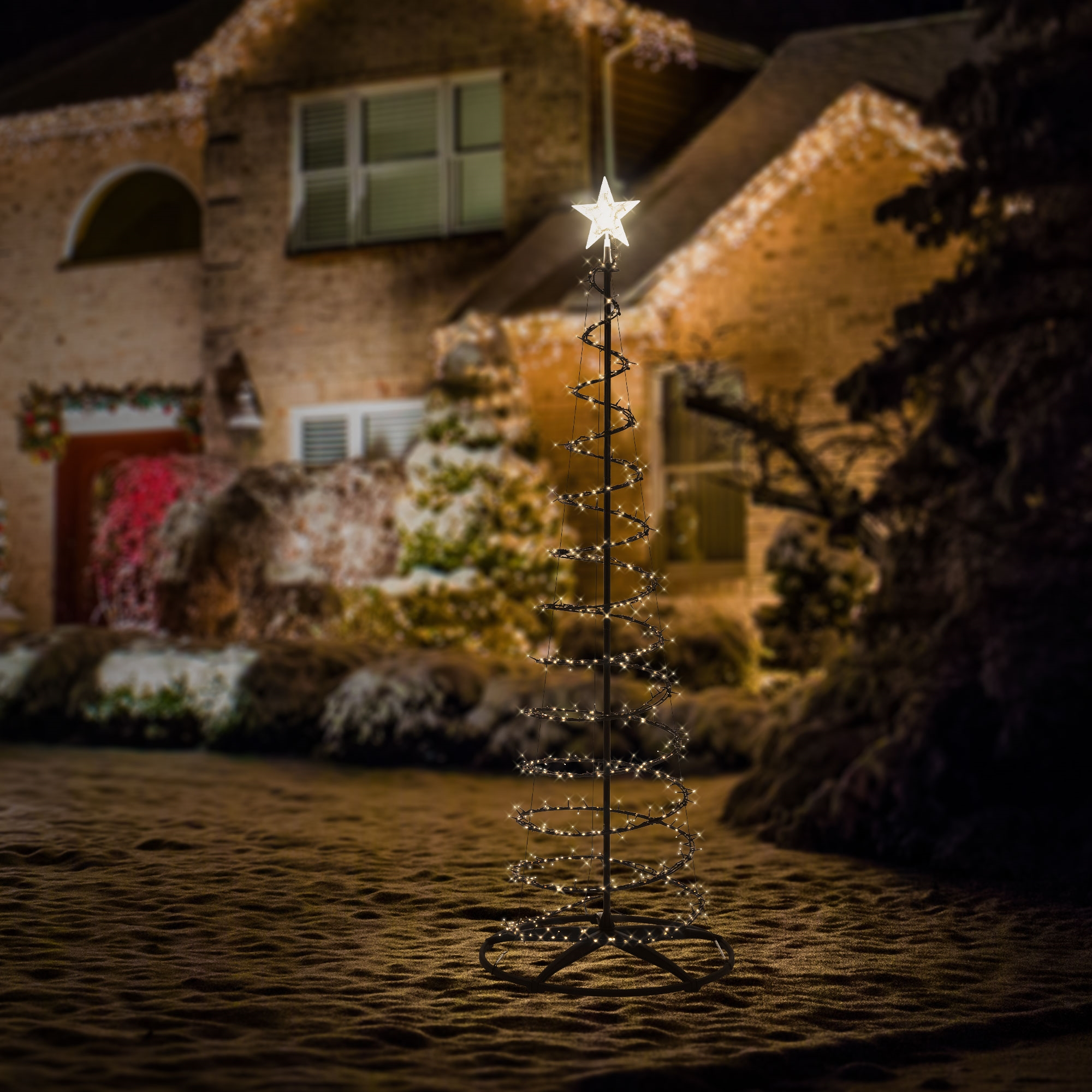 Ein spiralförmiger Weihnachtsbaum aus Lichterketten steht im Garten vor einem beleuchteten Haus.