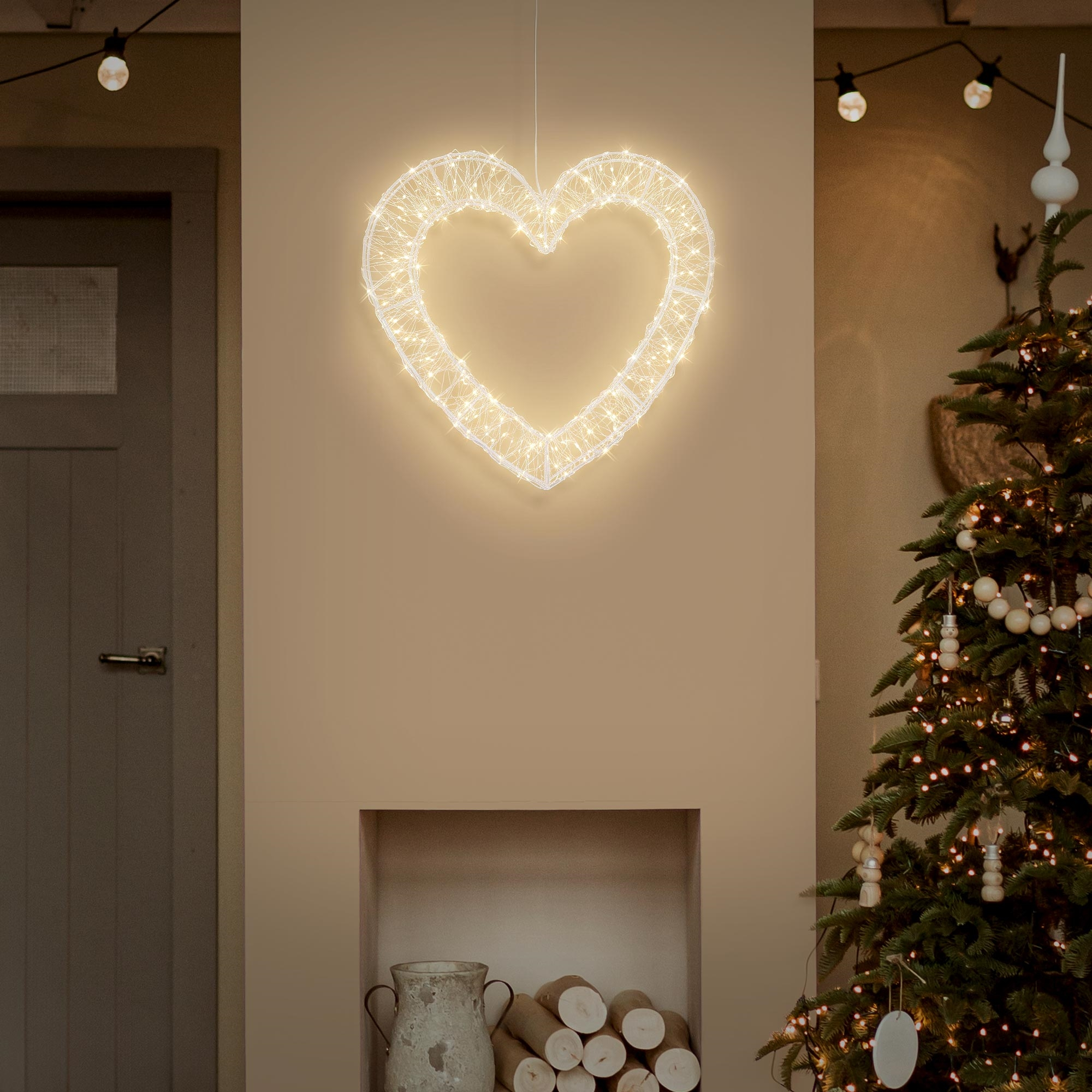 Ein leuchtendes Herz hängt an einer Wand über einem Kamin, daneben steht ein geschmückter Weihnachtsbaum.