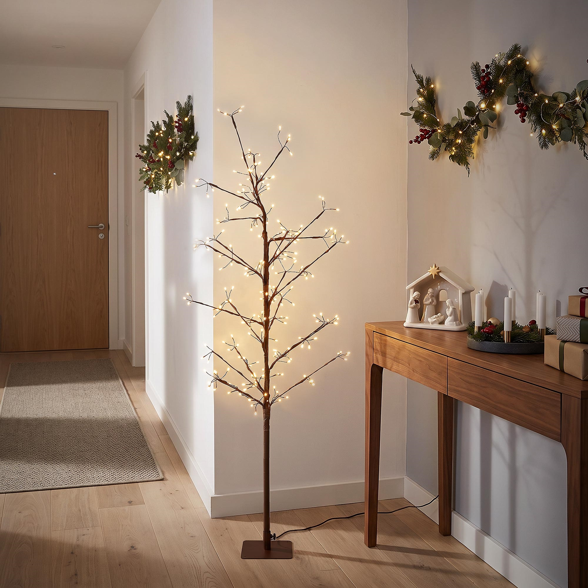 Ein beleuchteter künstlicher Baum steht in einem Flur neben einem Holztisch mit Weihnachtsdekoration.