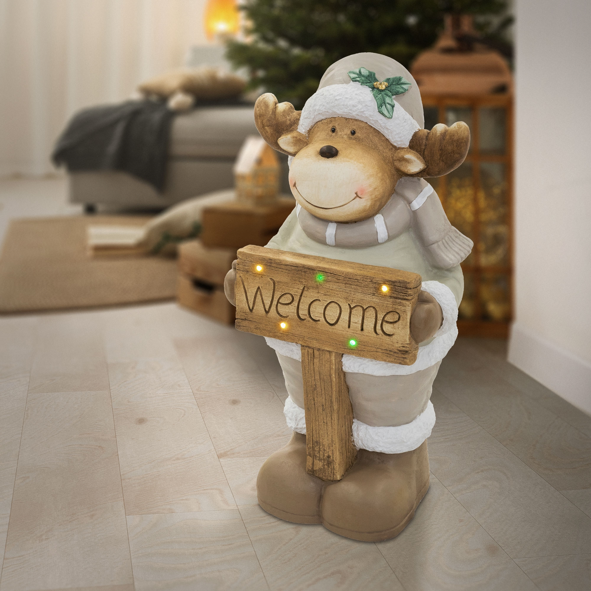 Eine Elchfigur im Winteroutfit mit Schal und Mütze hält ein beleuchtetes Willkommensschild.