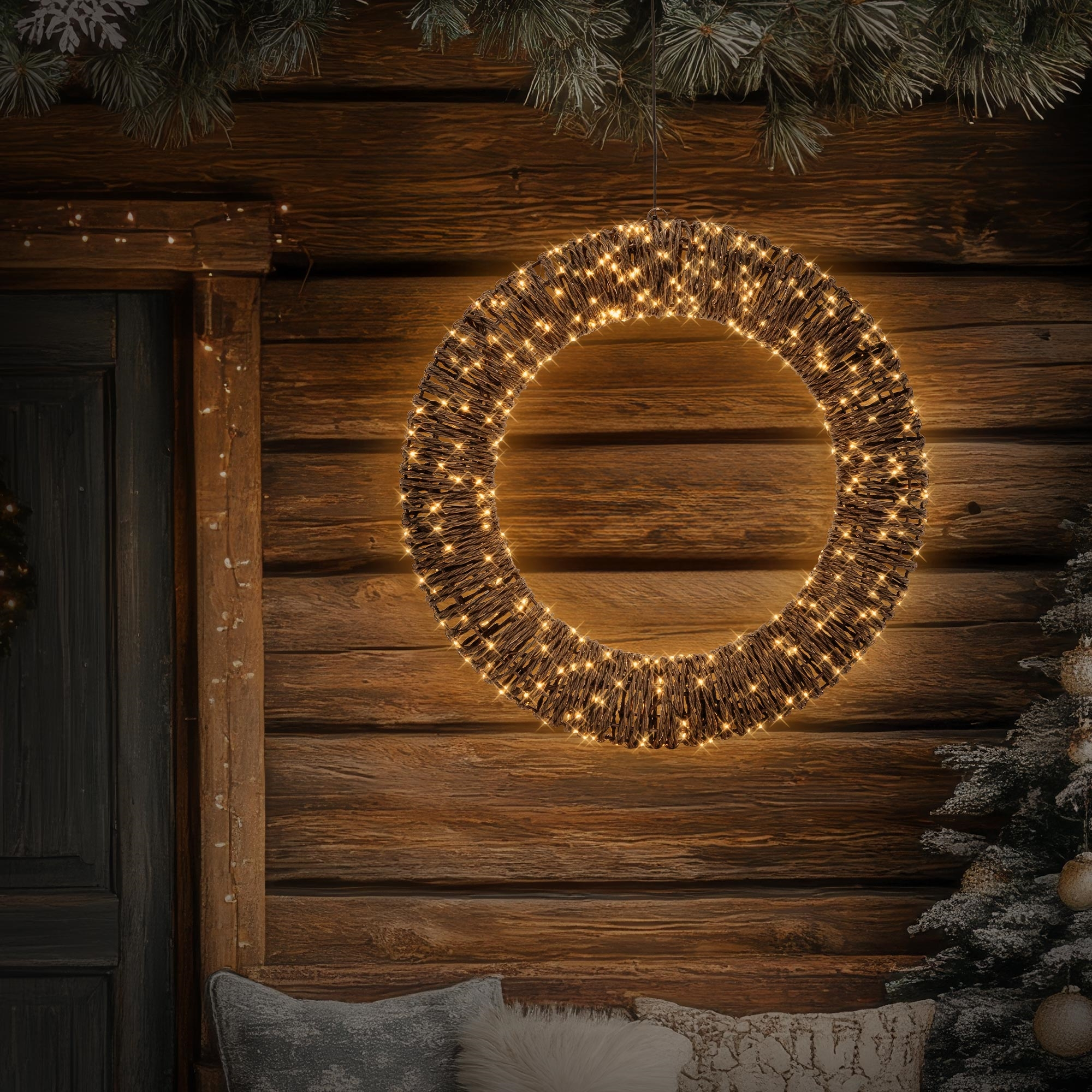 Beleuchteter Weihnachtskranz hängt vor einer dunklen Holzwand mit Tannengrün.