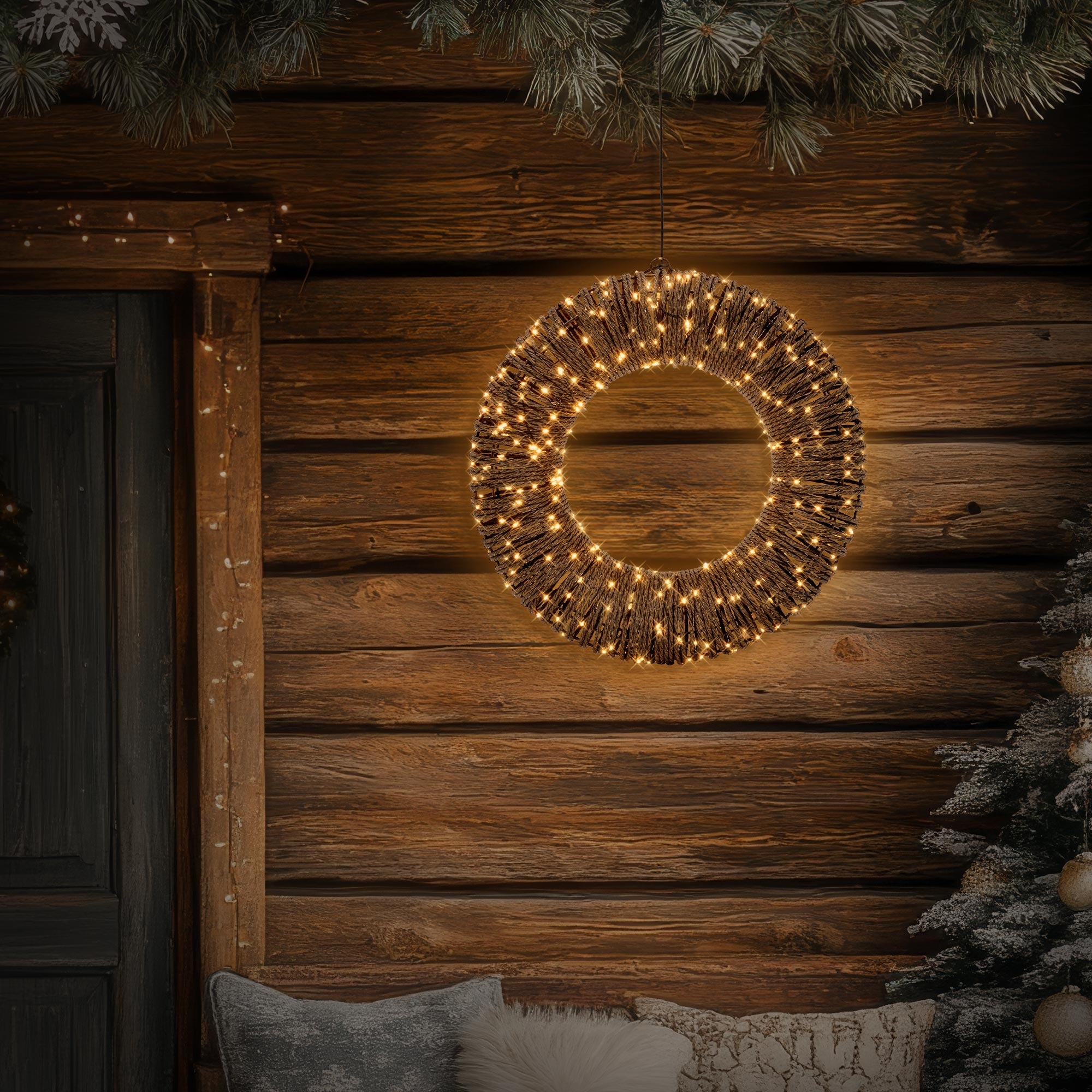 Beleuchteter Weihnachtskranz hängt an einer Holzwand mit Weihnachtsbaum und Kissen.