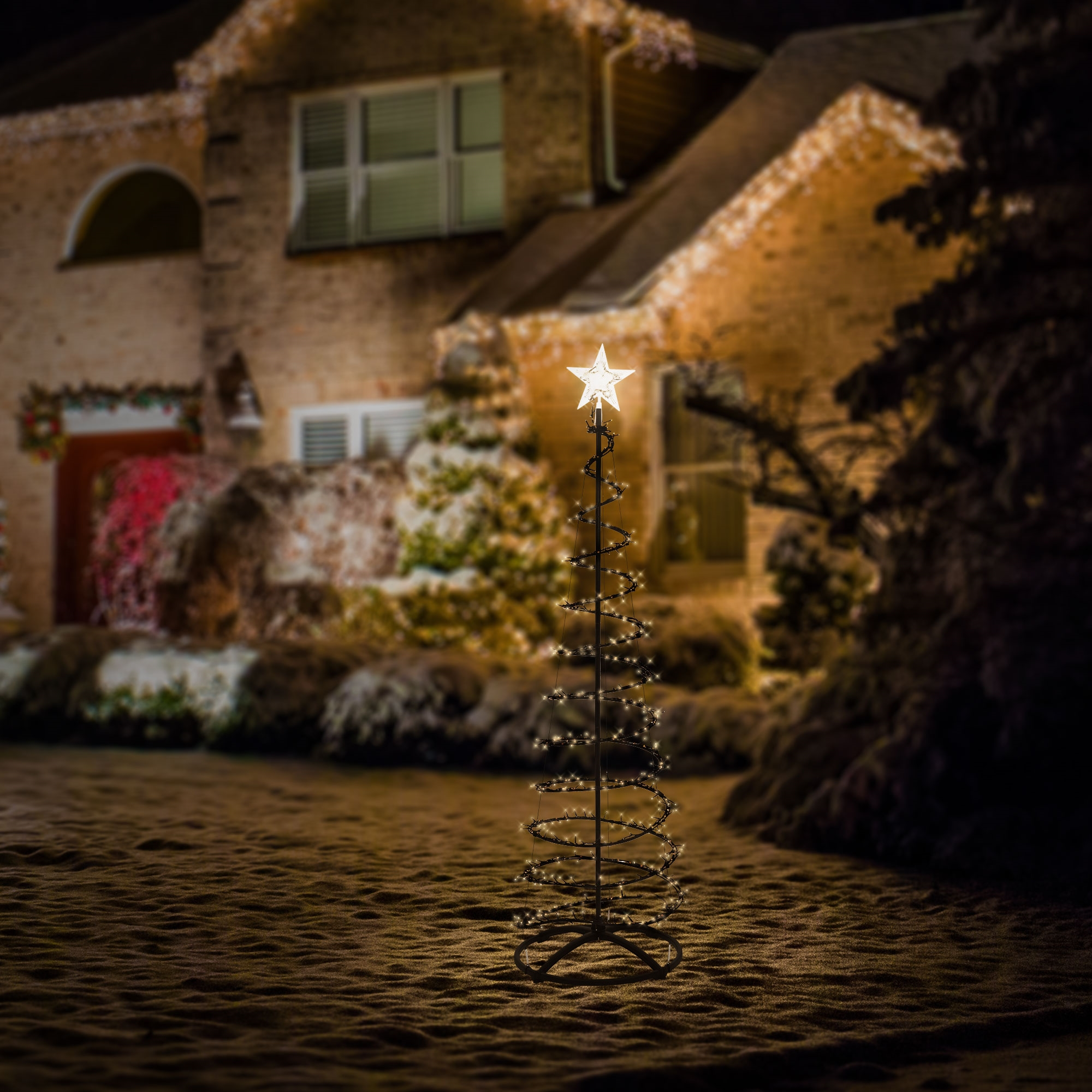 Ein leuchtender Spiral-Weihnachtsbaum mit Sternspitze steht nachts vor einem hell erleuchteten Haus.