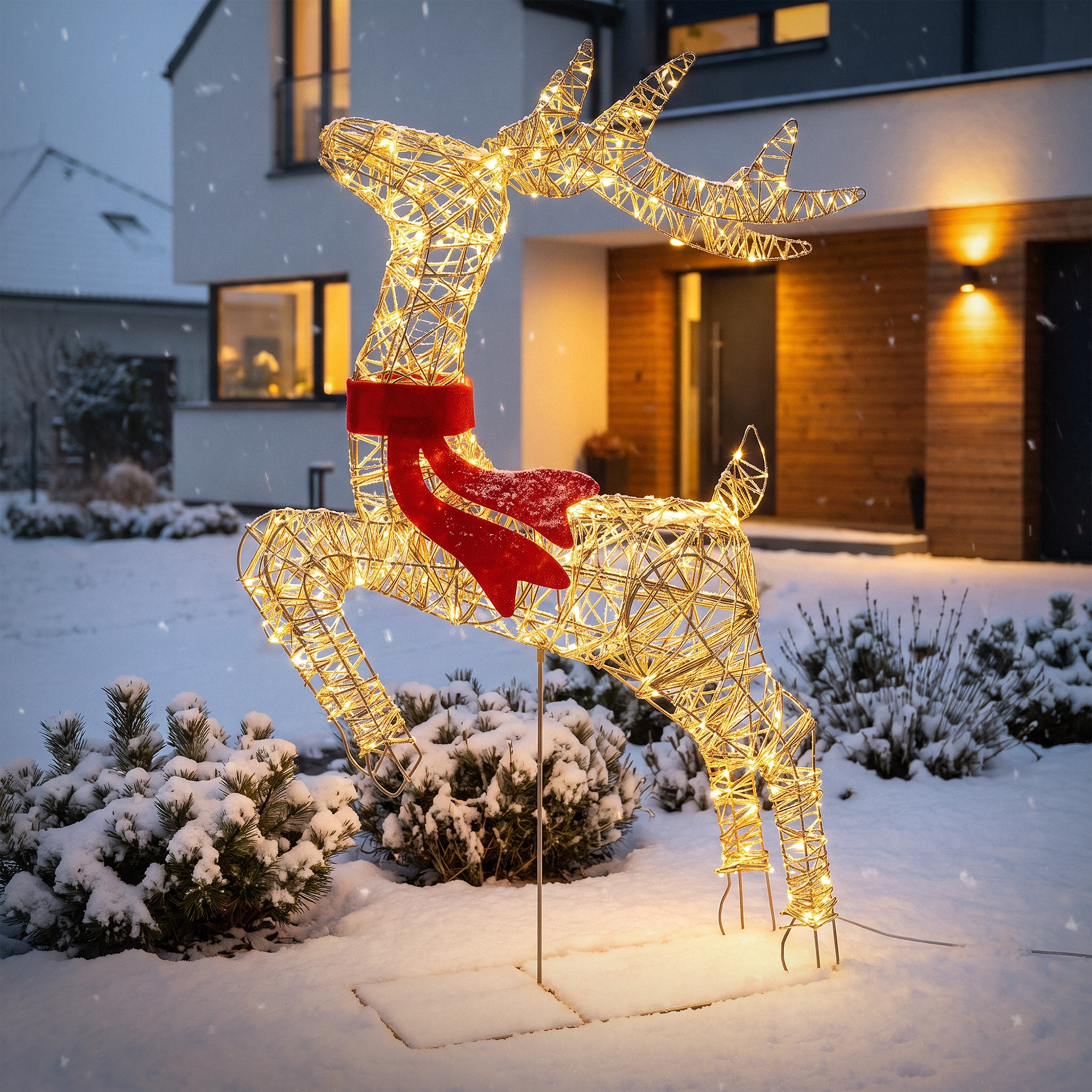 Beleuchtete Rentier-Skulptur mit rotem Schal im schnee bedeckten Garten vor einem modernen Haus.