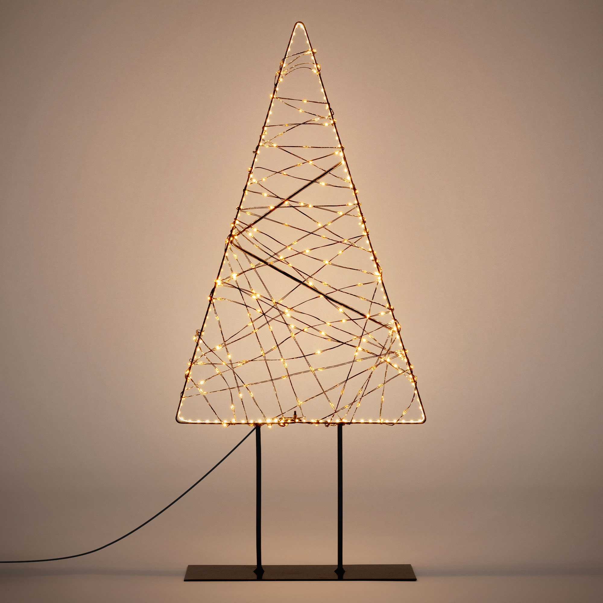 Eine Drahtmodell-Weihnachtsbaumskulptur mit leuchtenden Lichterketten steht auf einem schwarzen Sockel vor einer neutralen Wand.