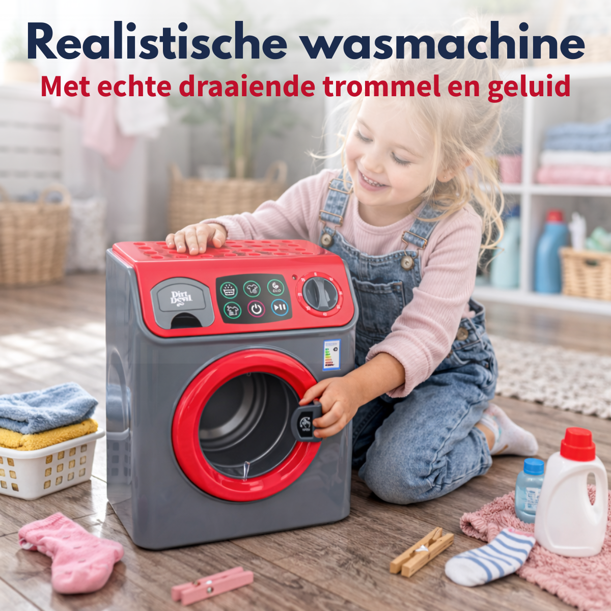 Une fillette souriante joue avec un lave-linge jouet gris et rouge sur un sol en bois.