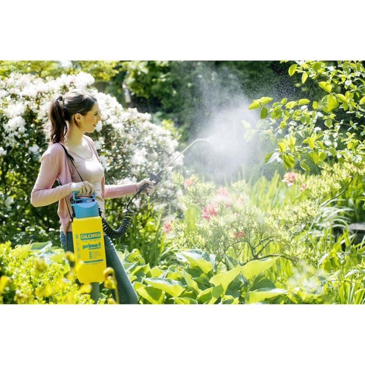 Frau sprüht mit einem gelben Gloria-Drucksprühgerät Wasser auf grüne Pflanzen in einem Garten.