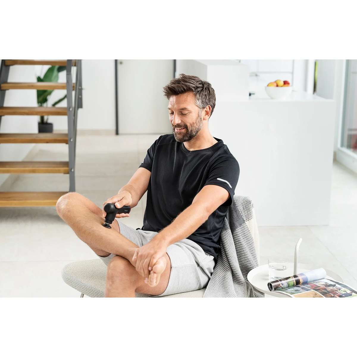 Un uomo seduto su una sedia in una stanza luminosa massaggia la gamba con un piccolo dispositivo nero.